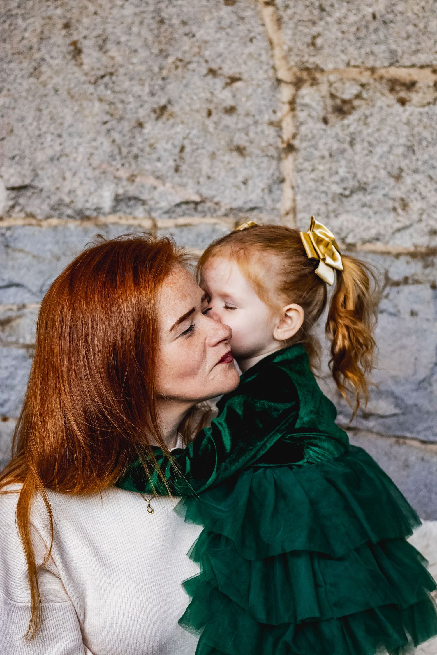 A tender moment shared between parent and child wearing white and emerald green velvet dress against stone wall.