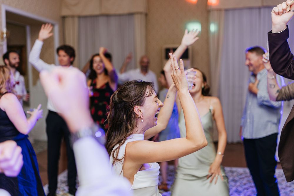 Wedding guests dance and celebrate on a crowded dance floor with colorful lighting and raised hands in the air.