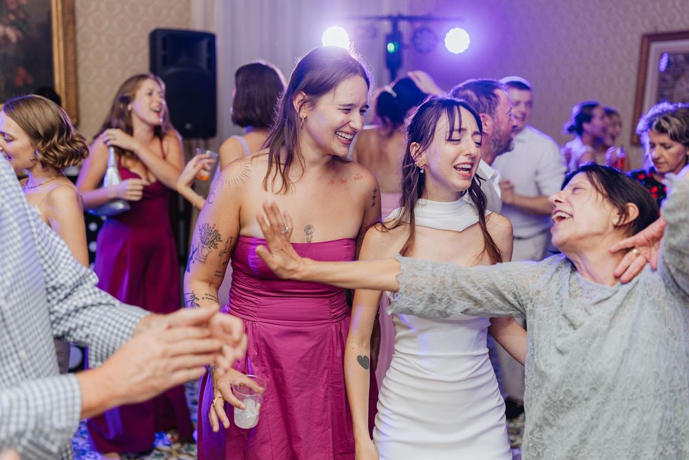 Wedding guests dance and celebrate on a crowded dance floor illuminated by purple lighting at a reception party.