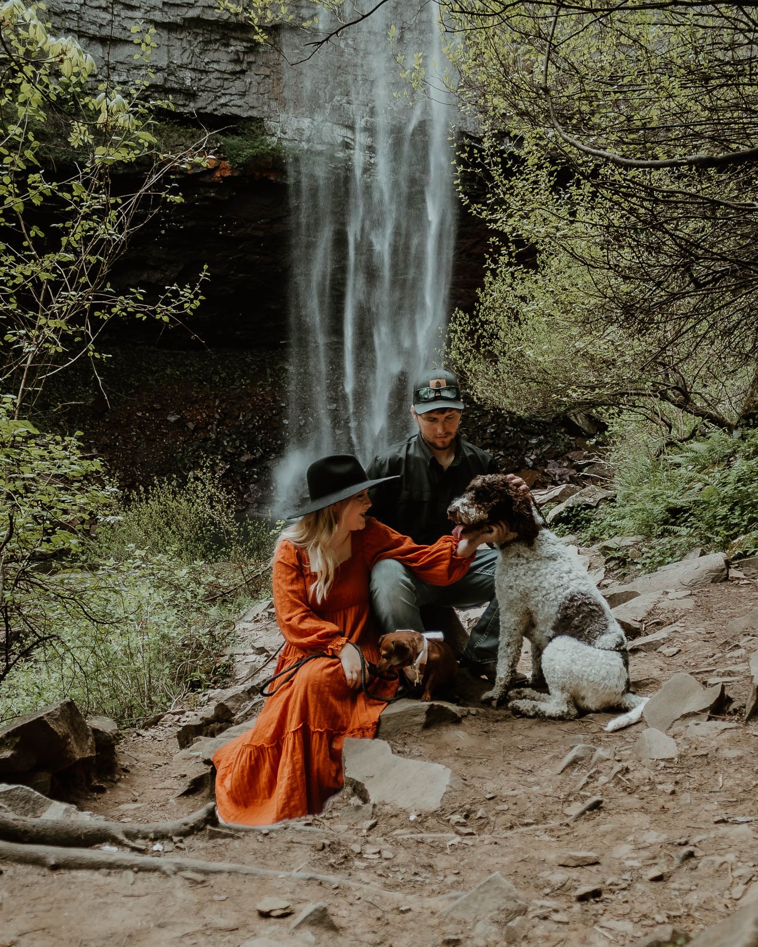 Couple sitting with their dog by a scenic waterfall on a hiking trail.