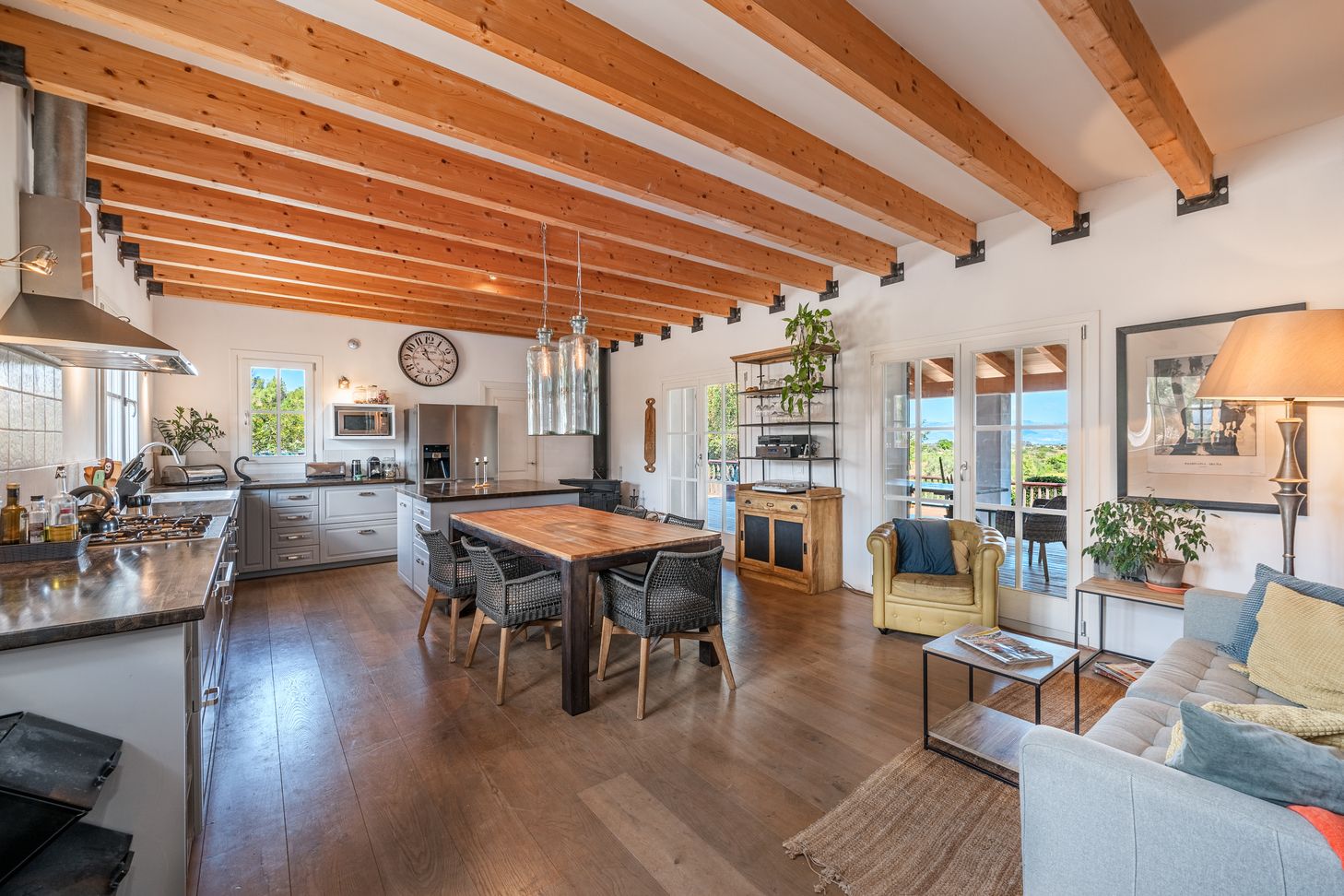 Modern open concept living space with exposed wooden beams, white walls, and natural light flowing through large windows.