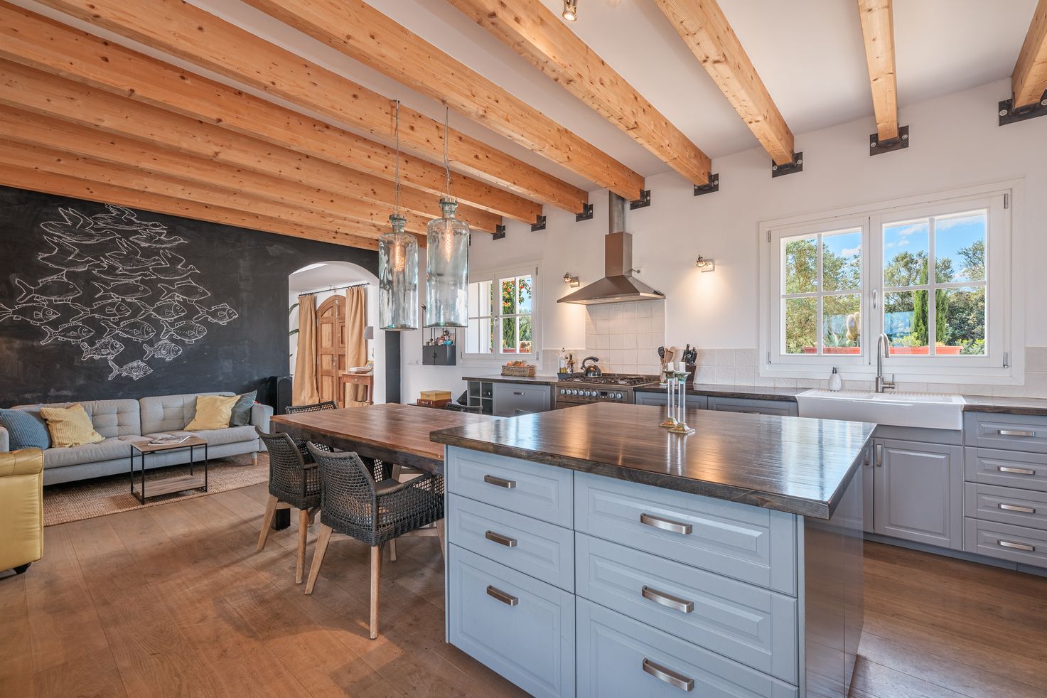 Modern kitchen with exposed wood beams features gray cabinets, stainless appliances, and large windows overlooking greenery.