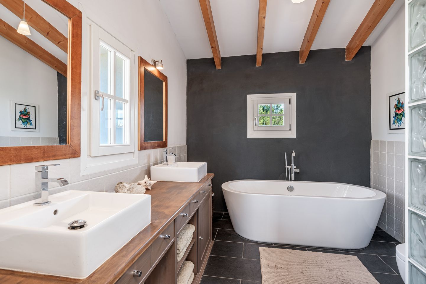 Modern bathroom interior with freestanding white tub, dark accent wall, exposed wooden beams, and double vessel sinks.
