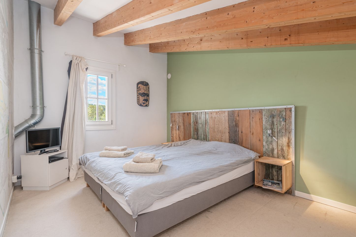 Modern bedroom with rustic wood headboard, green accent wall, exposed wooden beams, and natural lighting from window.