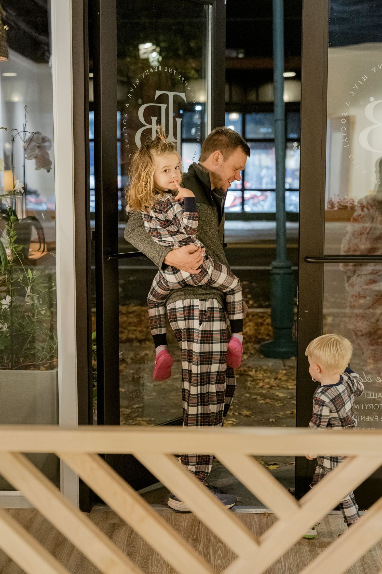 Two children in matching plaid christmas pajamas look through glass windows of a building at night.