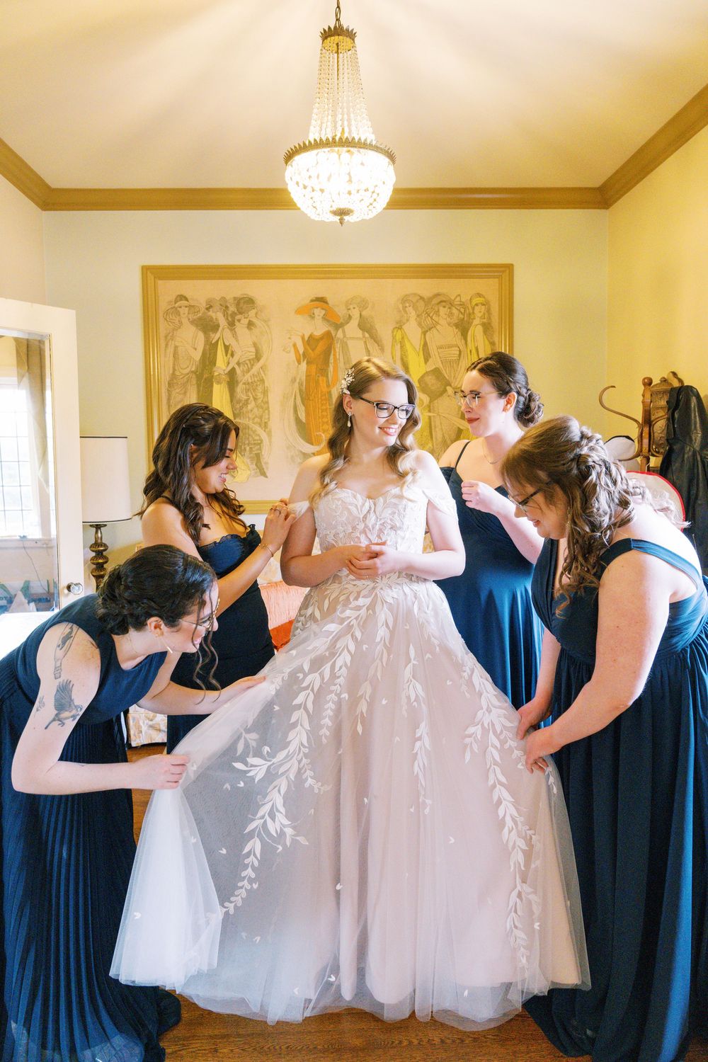 Bridesmaids in navy blue dresses help arrange a white wedding gown in an elegant room.