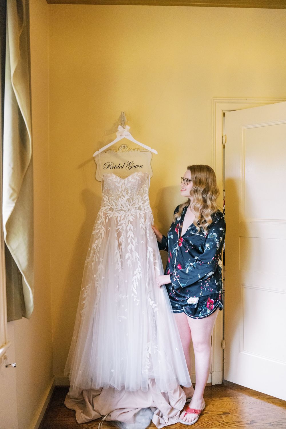 A white lace wedding gown hangs on display in a room with yellow walls.