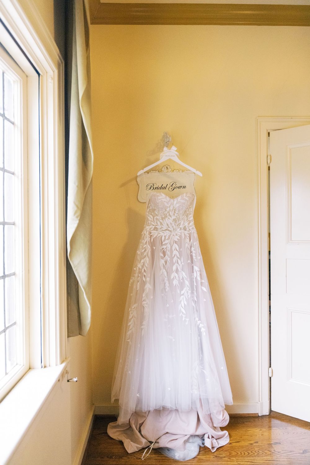 White lace wedding dress hangs elegantly against cream colored wall near window.