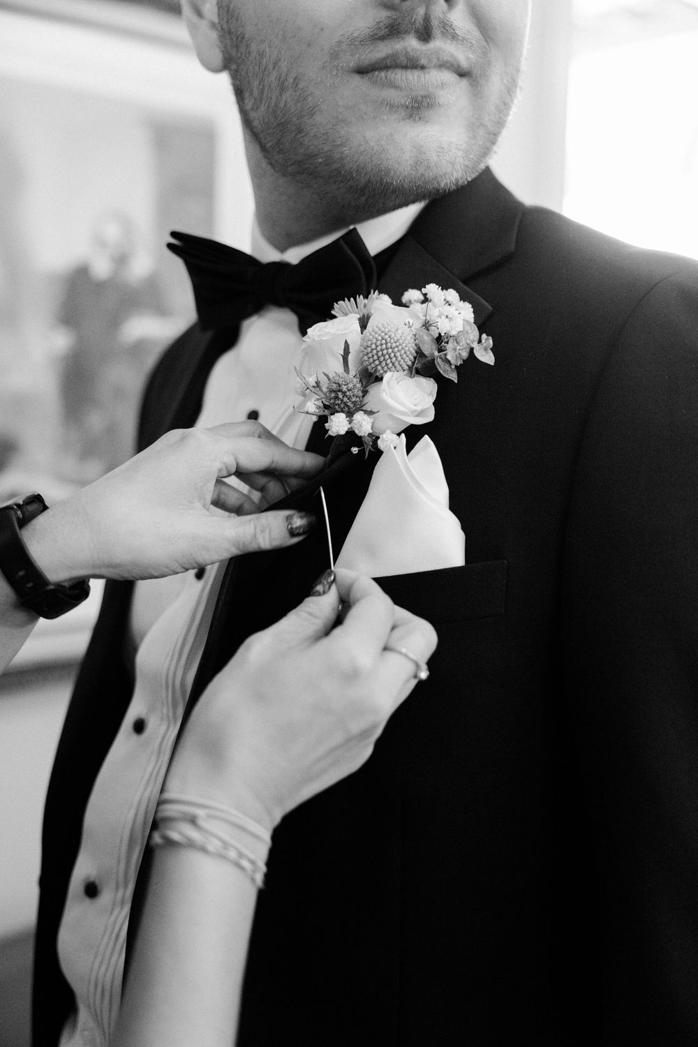 Close-up of hands pinning a white boutonniere onto a black tuxedo lapel.