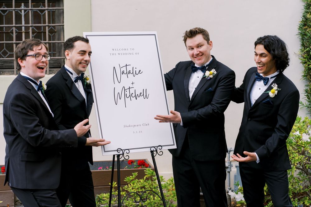 Groomsmen in black tuxedos gather around a wedding welcome sign displayed on an easel.