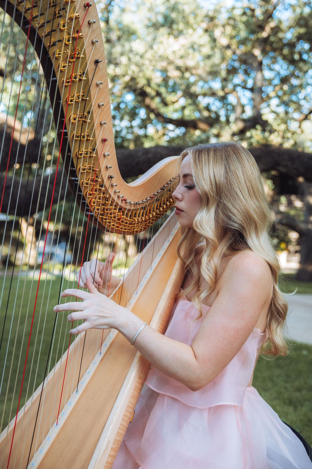 Musician in pink dress performing with a golden harp outdoors among trees.