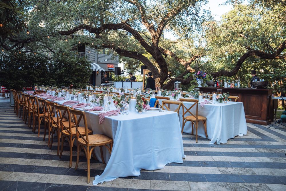 Outdoor wedding reception setup with white tablecloths and wooden chairs under trees.