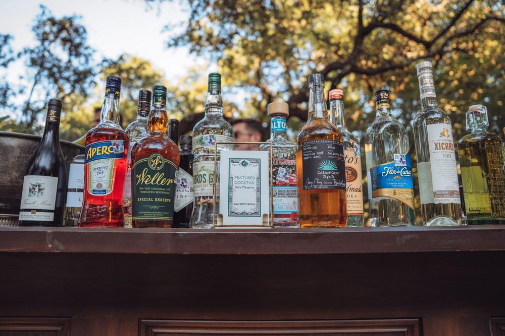 Collection of liquor bottles displayed on wooden bar shelf at outdoor event.