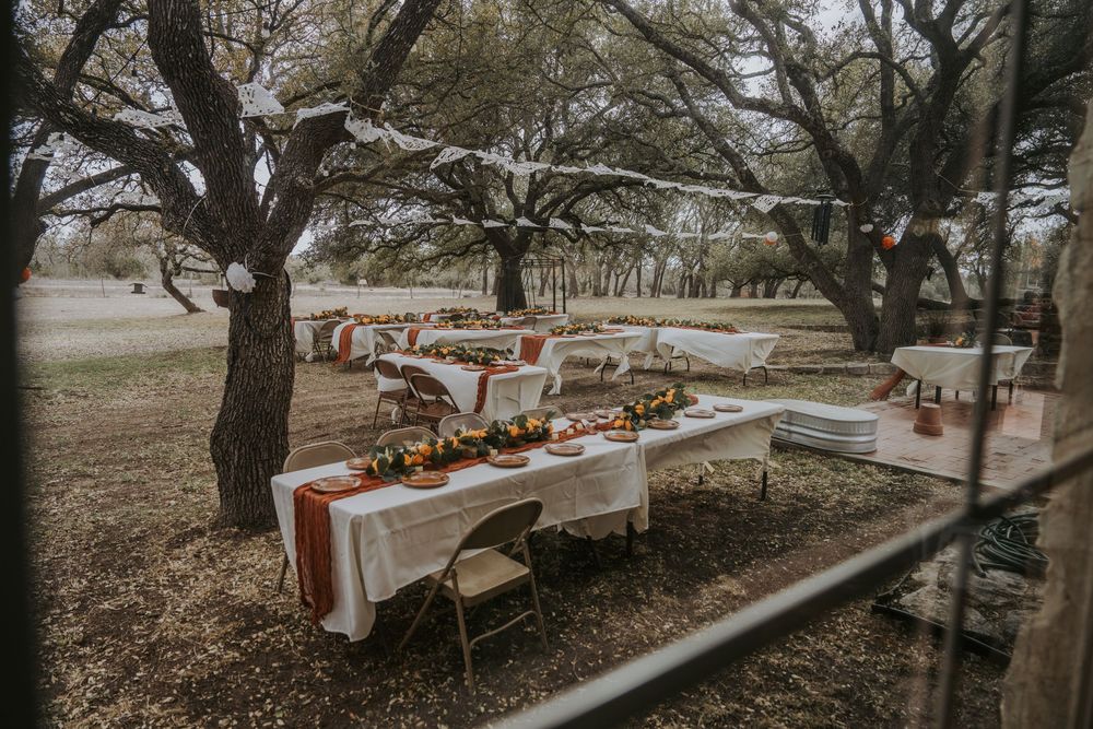 Rustic outdoor wedding reception setup with wooden tables and chairs under trees at dusk.