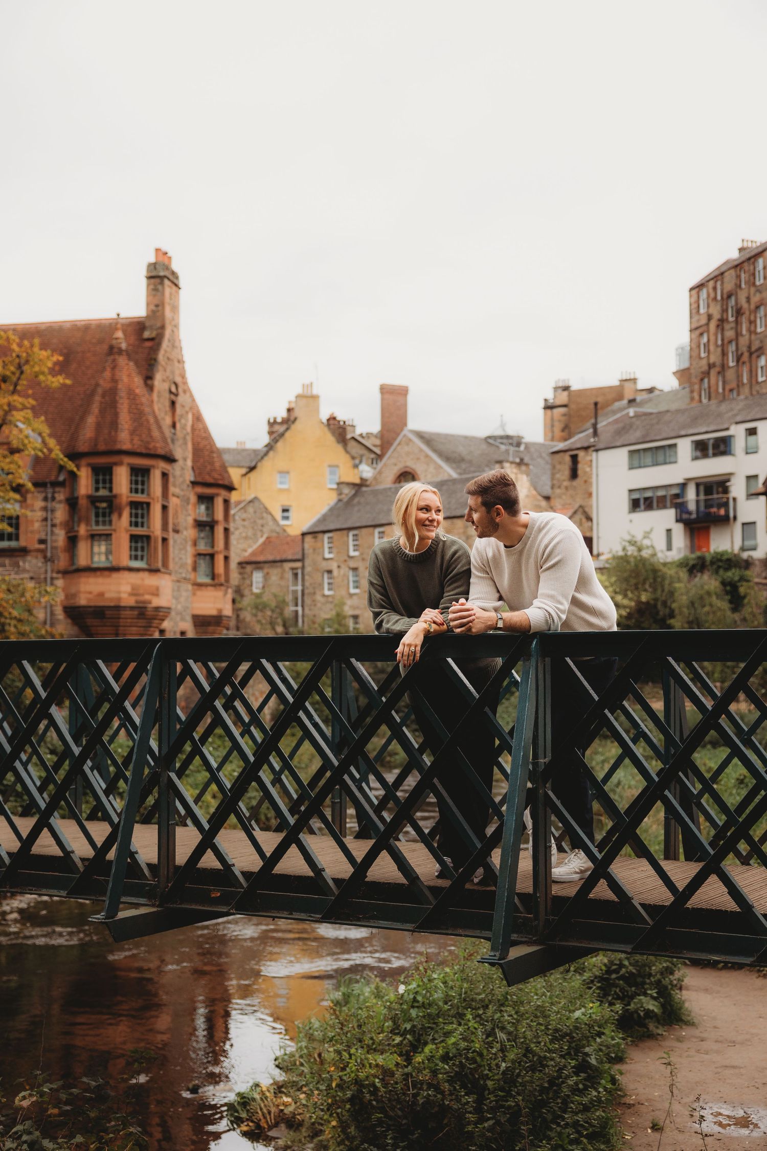 Couple sitting on a metal bridge railing overlooking a river with historic stone buildings in the background. Dean Village, Edinburgh