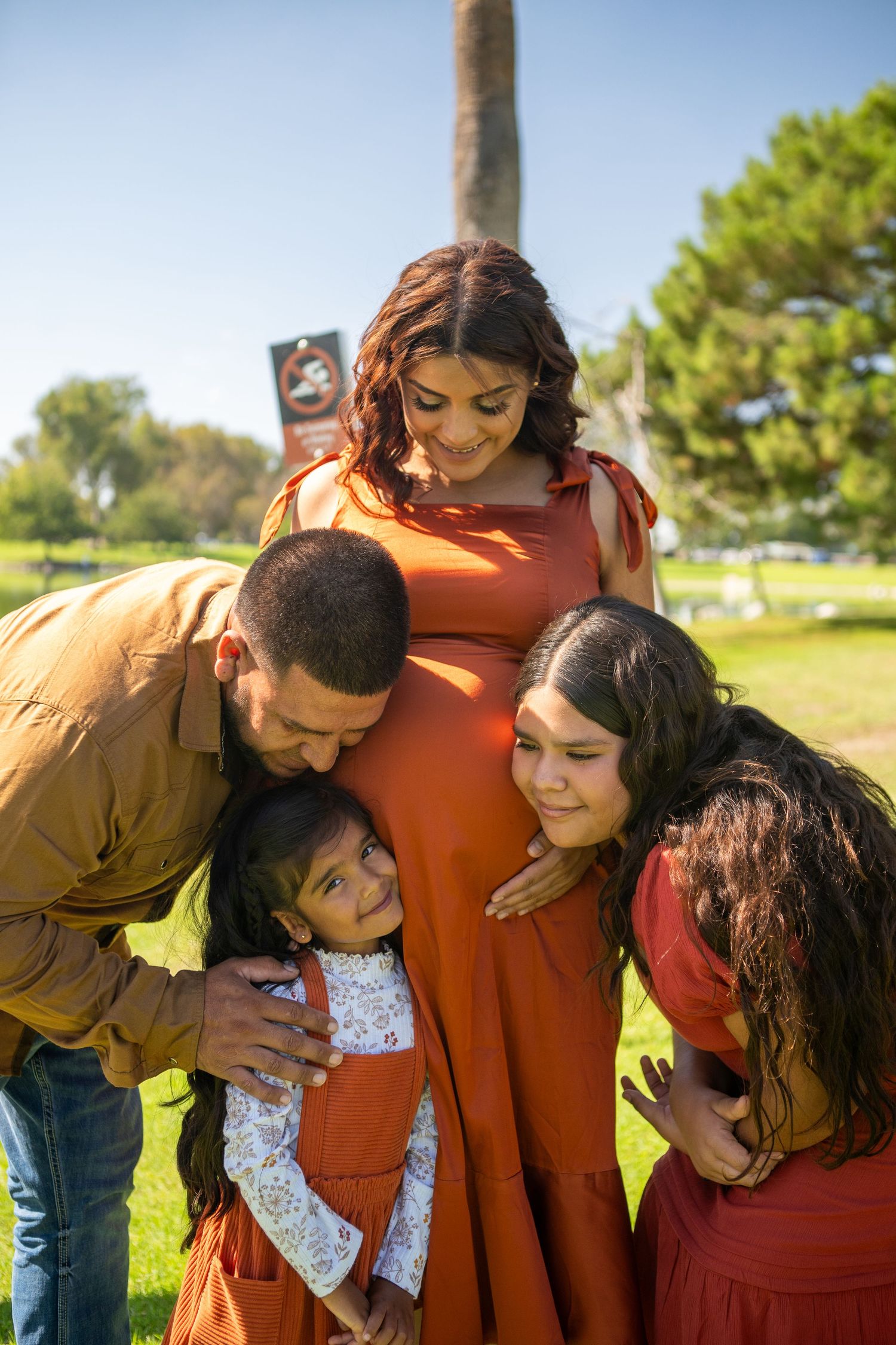Family shares intimate moments together in a park setting with warm sunlight and trees in the background.