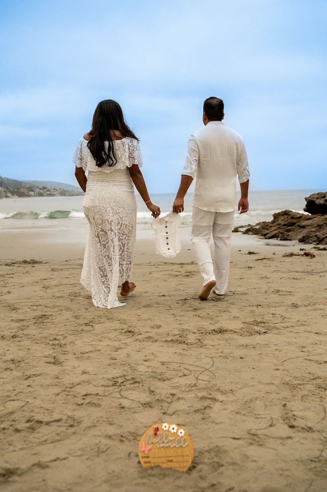 Couple in white walking away on sandy beach holding hands.