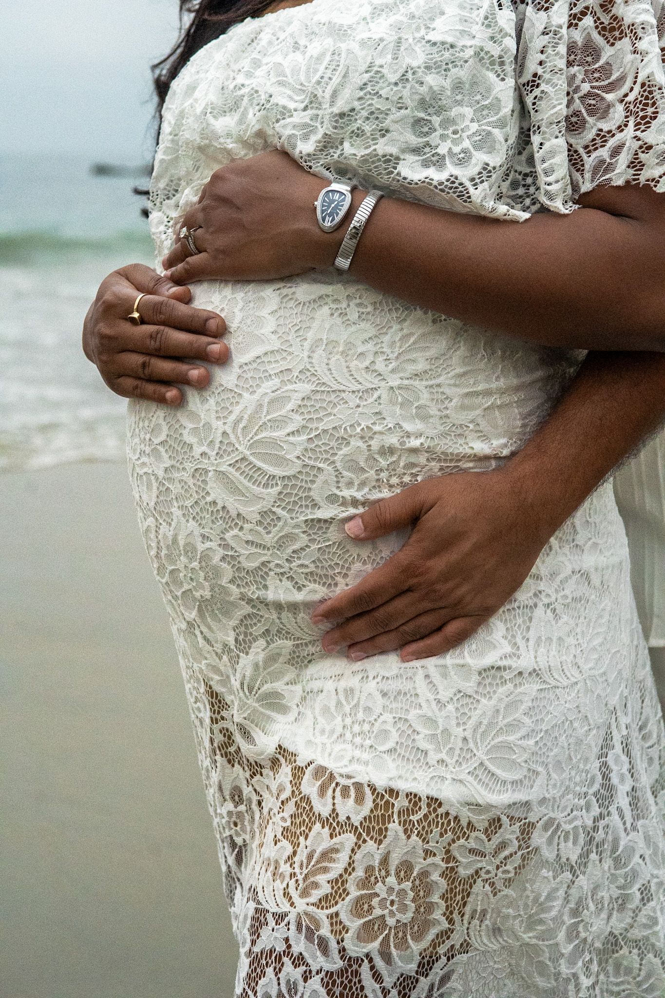 Close-up of hands cradling a pregnant belly in white lace dress against beach backdrop.