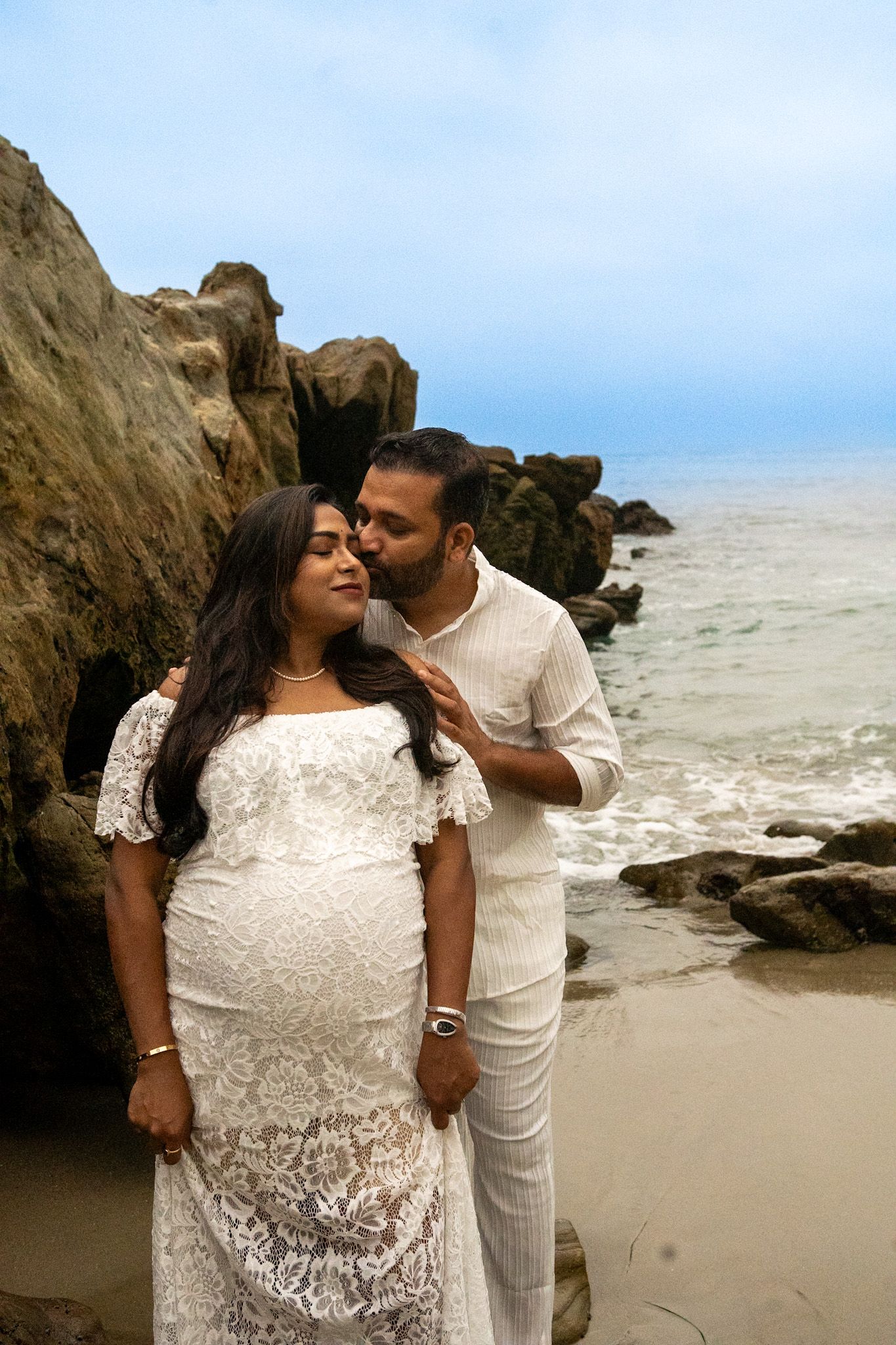 Maternity photoshoot on beach with rocky cliffs, couple wearing white outfits.