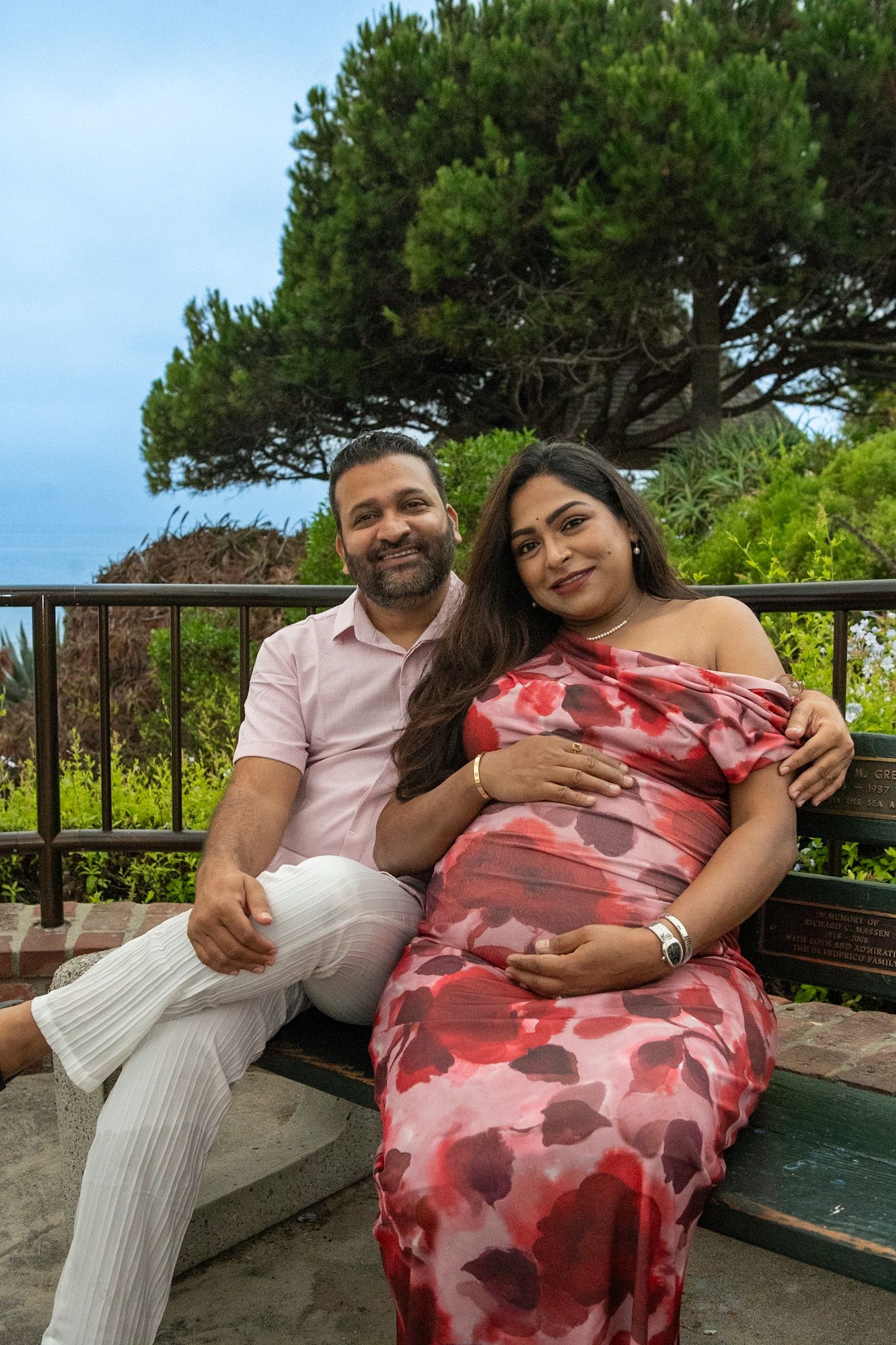 Couple relaxes on bench during coastal maternity photoshoot.