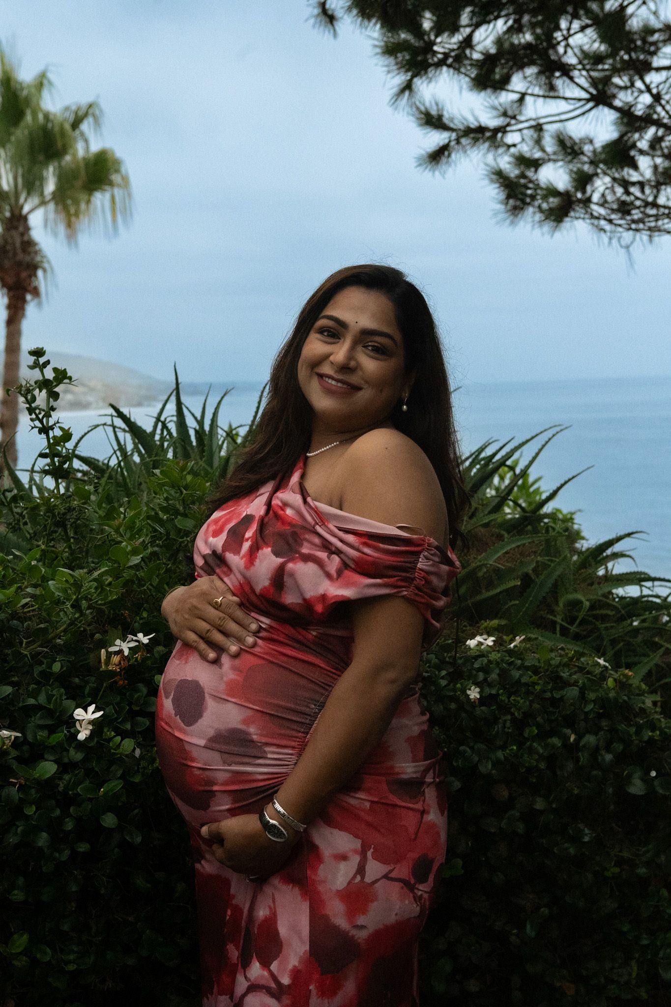 Expectant mother in red floral dress poses in tropical garden setting.