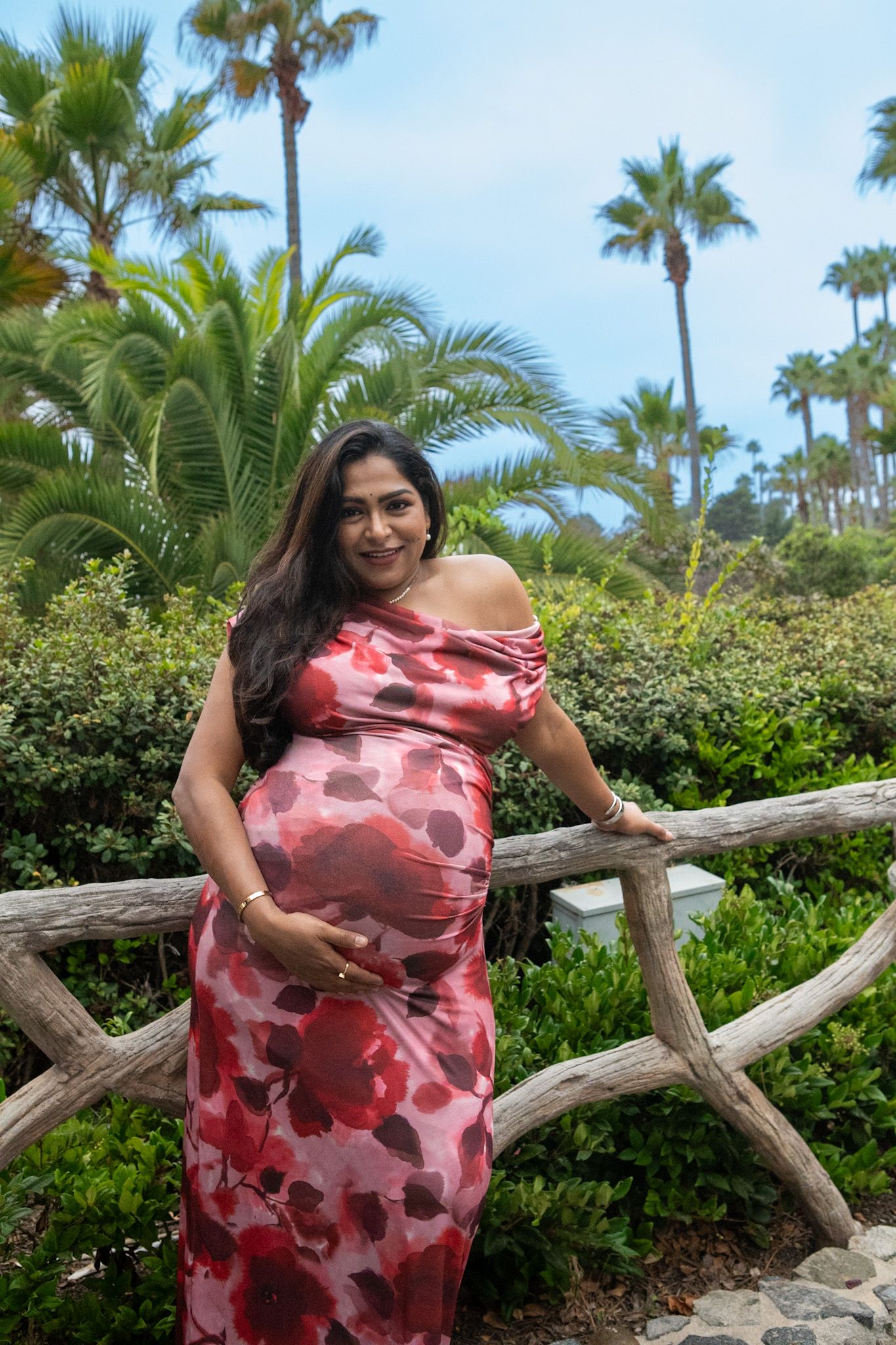Pregnant woman in red floral dress poses by palm trees in tropical garden.