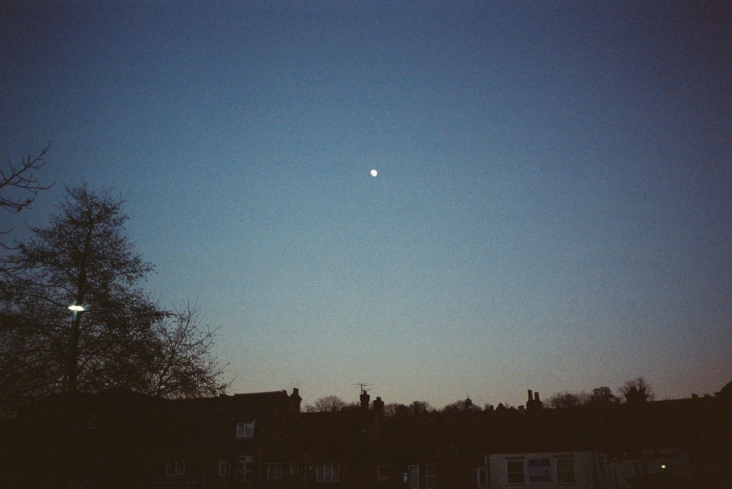 film photo of the moon in the sky at dusk, over silhouetted rooftops in an english village.