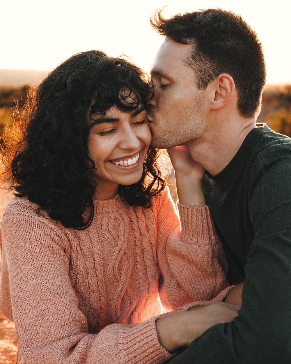 Man kissing smiling woman on cheek during warm sunset engagement photo session.