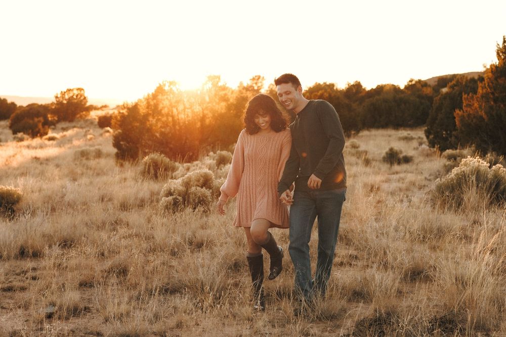 Happy couple walking hand in hand through golden meadow at sunset, smiling warmly.