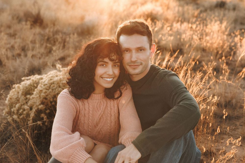 Couple smiling together in golden field at sunset, woman in pink sweater leaning on man in green shirt.