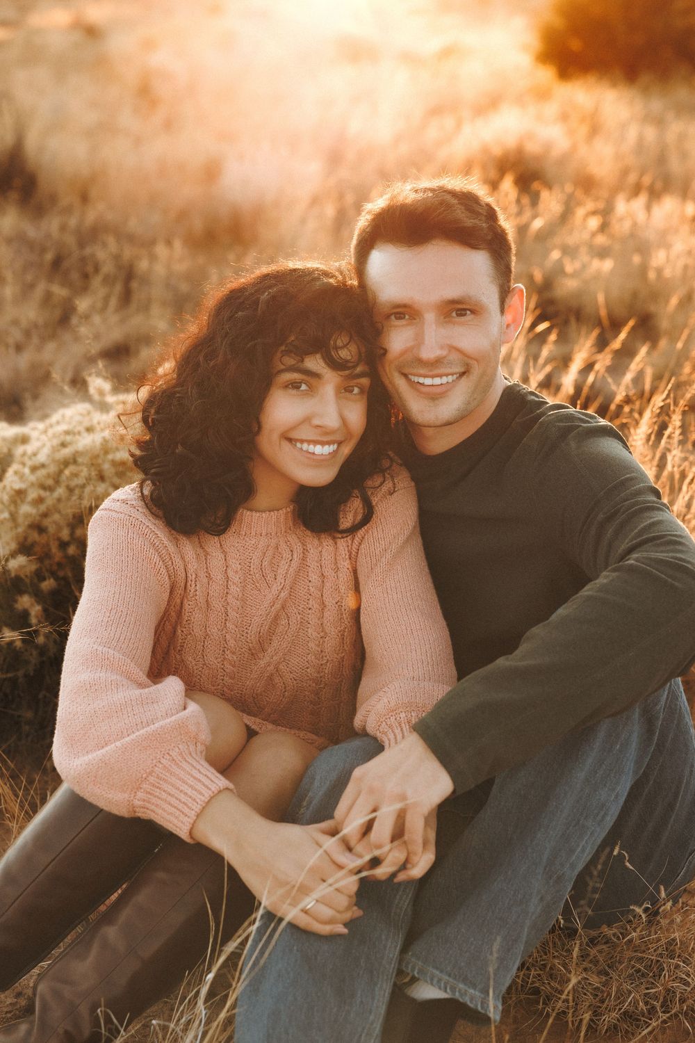 Happy couple sitting in autumn field holding hands, bathed in warm golden sunset light.
