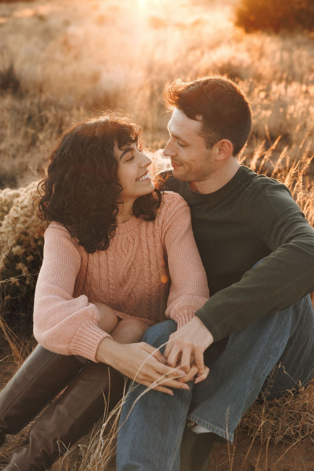 Romantic couple gazing at each other lovingly while sitting in sunlit golden grass field.
