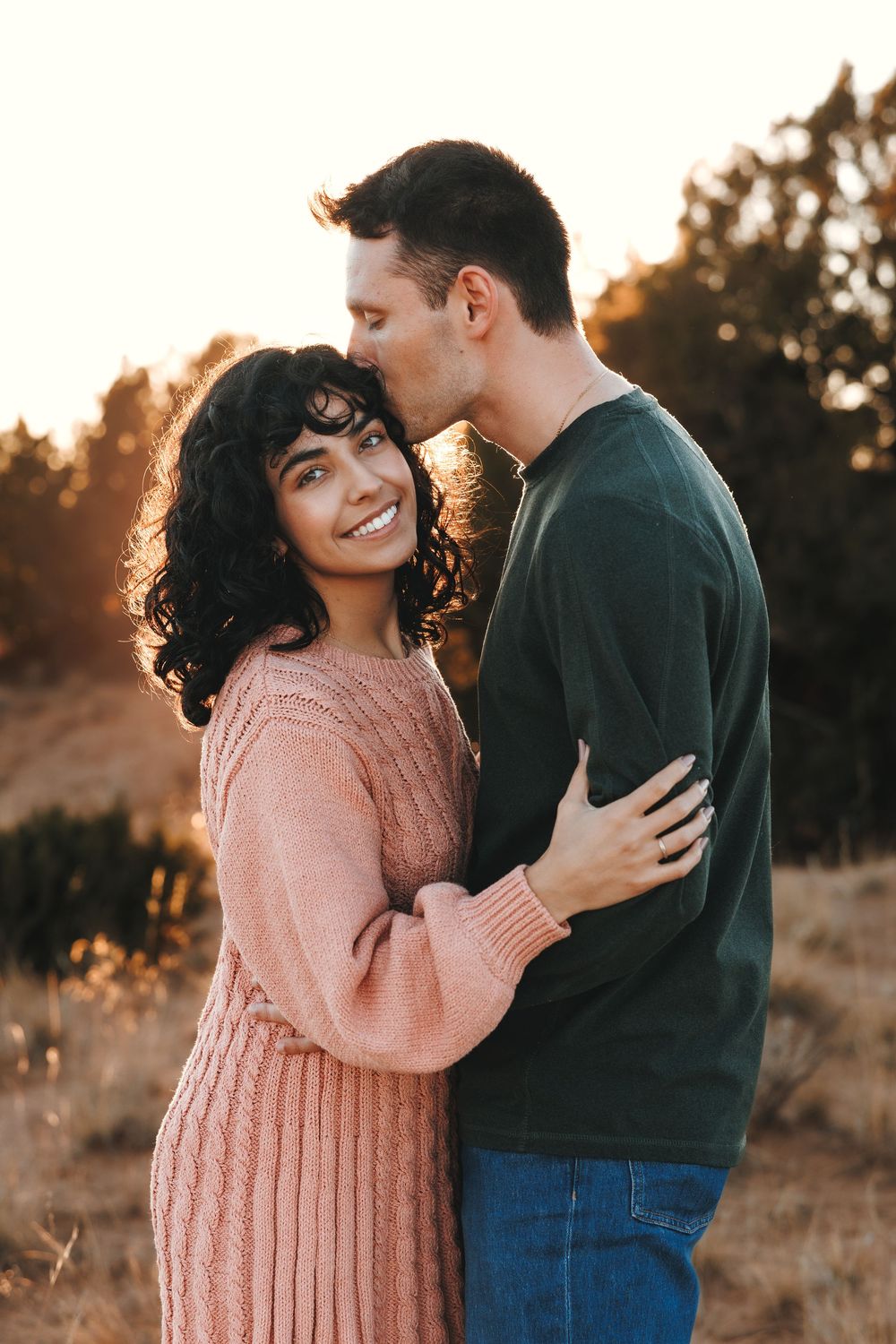 Man kissing woman on temple during golden hour outdoor couples photo session.