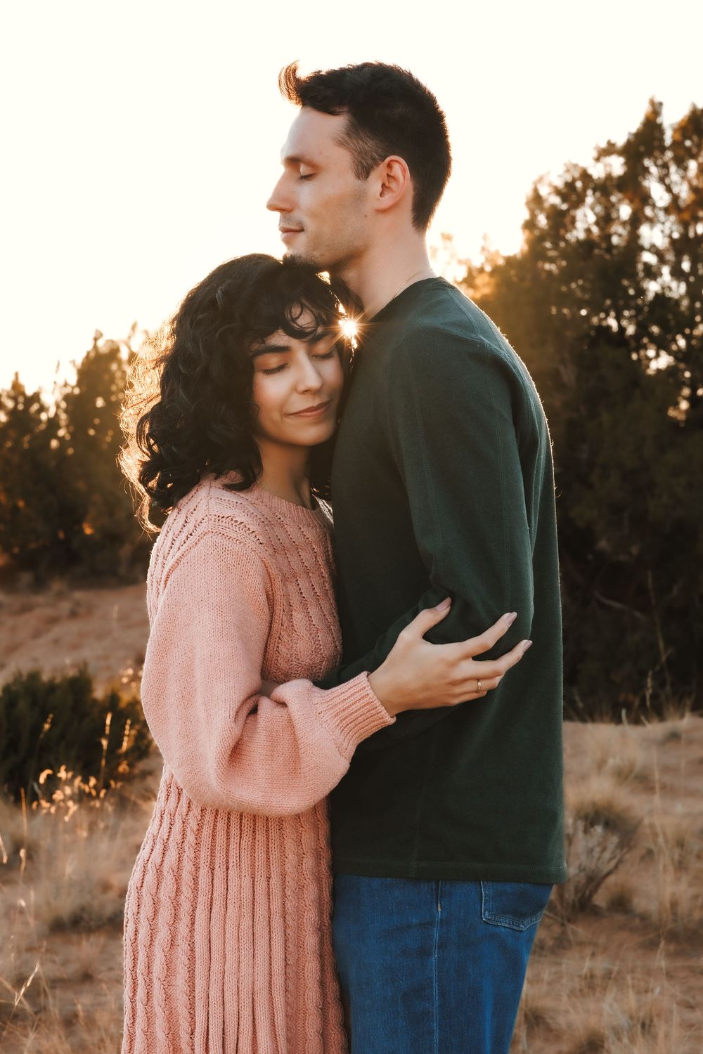 Couple embracing at sunset in desert field, woman in pink sweater resting on mans chest.