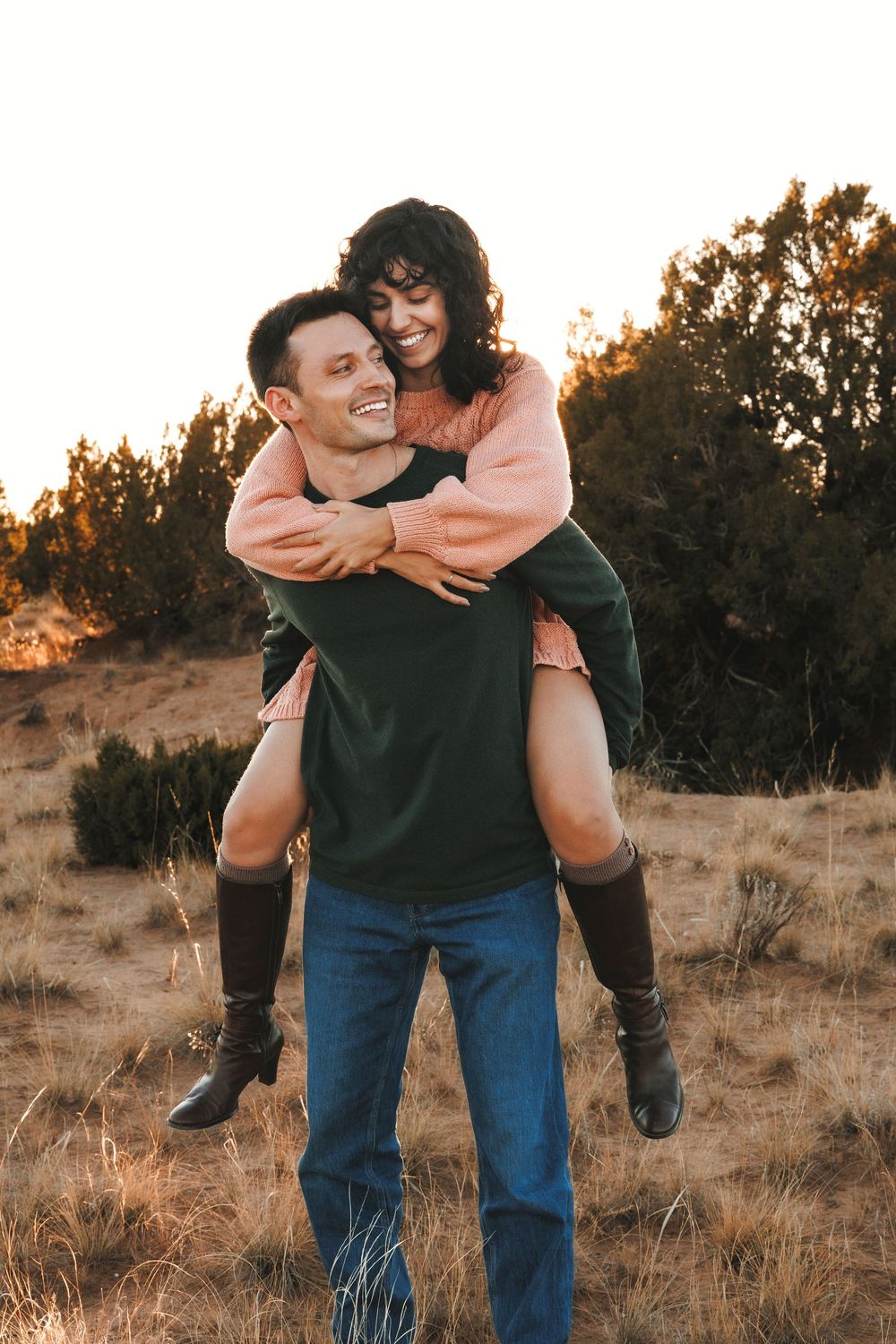 Man giving woman a piggyback ride in golden field at sunset, both smiling joyfully.