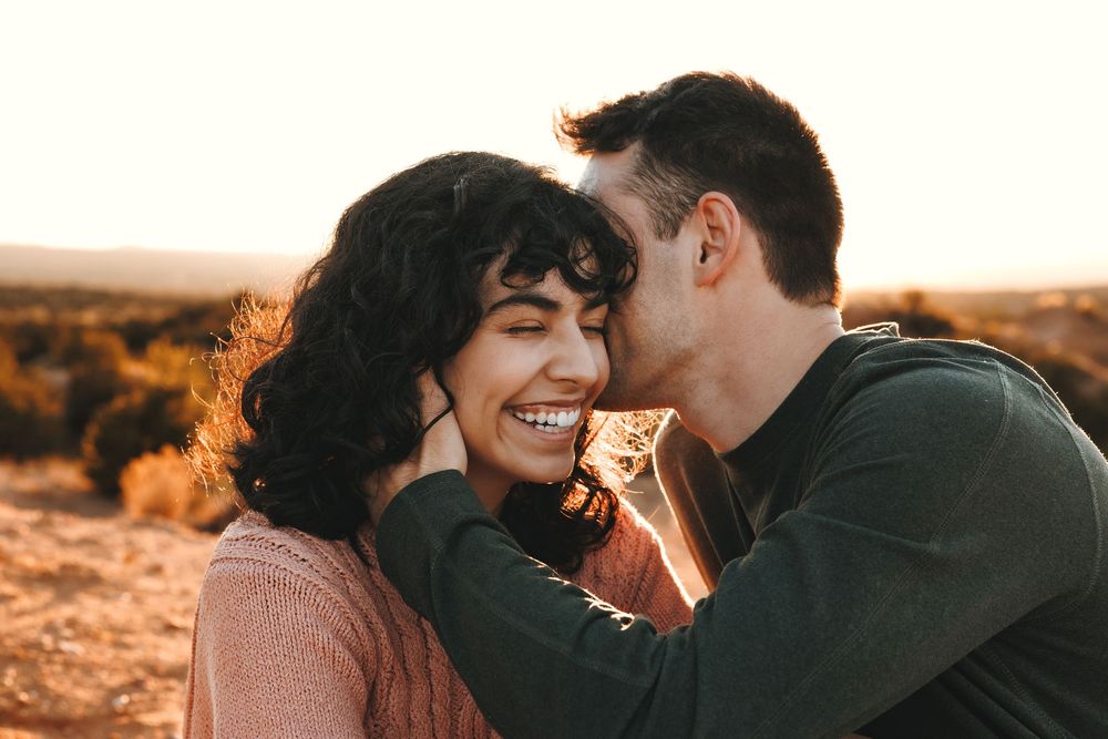 Couple sharing a tender cheek kiss moment outdoors at warm sunset, both smiling.