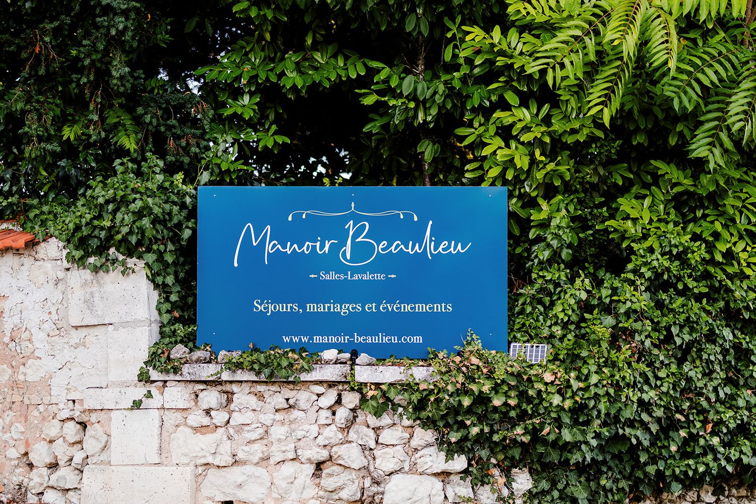 Blue sign for Manoir Beaulieu mounted on an ivy-covered stone wall.