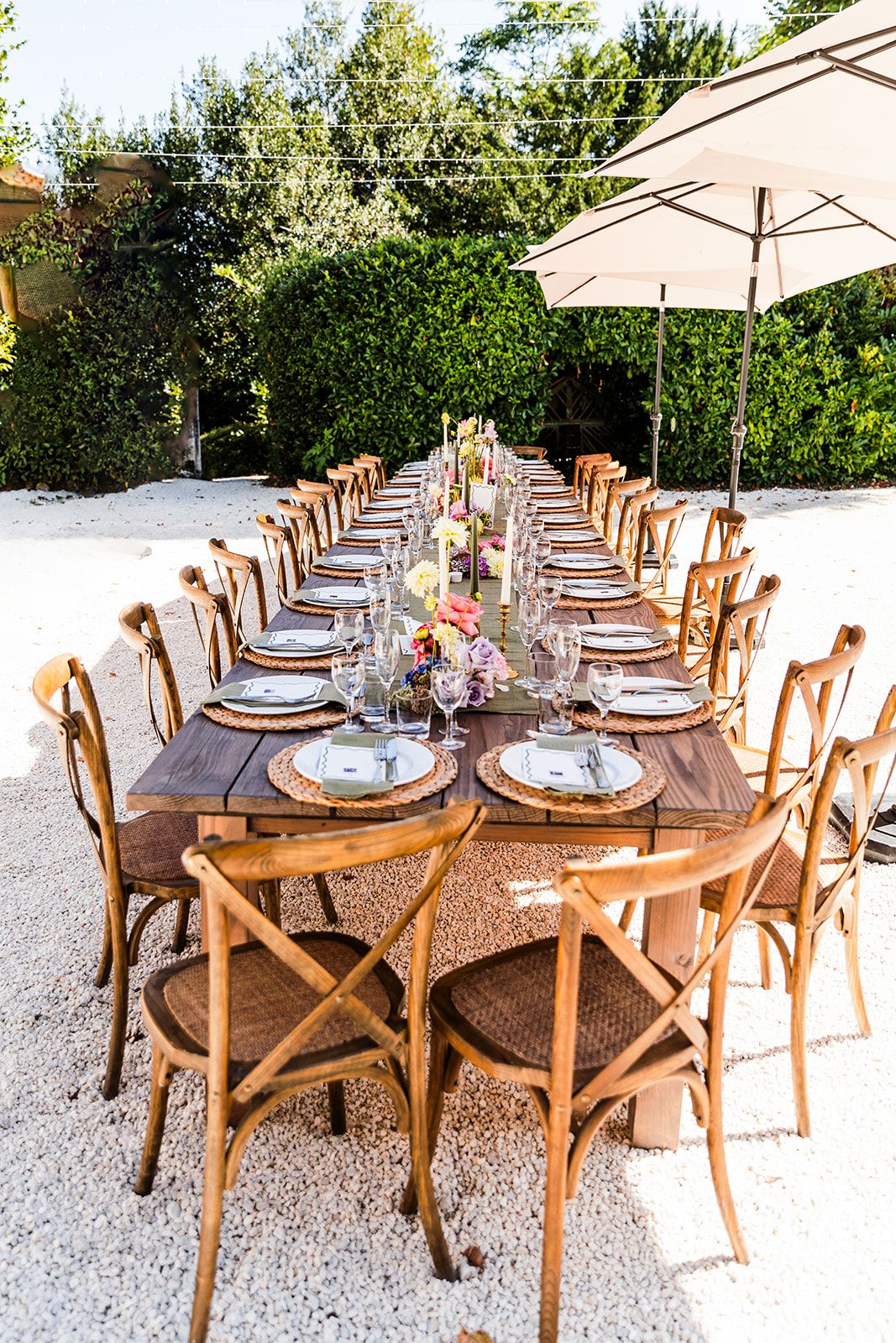 Long wooden dining table set with white plates and cross-back chairs under outdoor umbrellas on gravel patio.