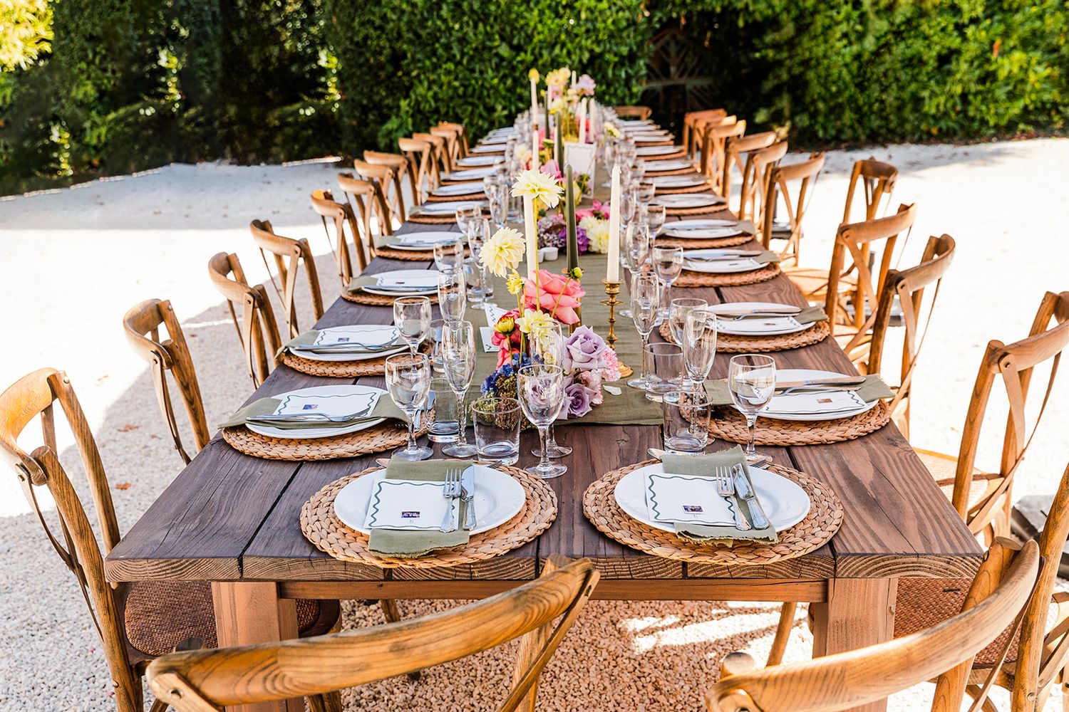 Long wooden banquet tables set with elegant place settings for an outdoor wedding reception under natural sunlight.