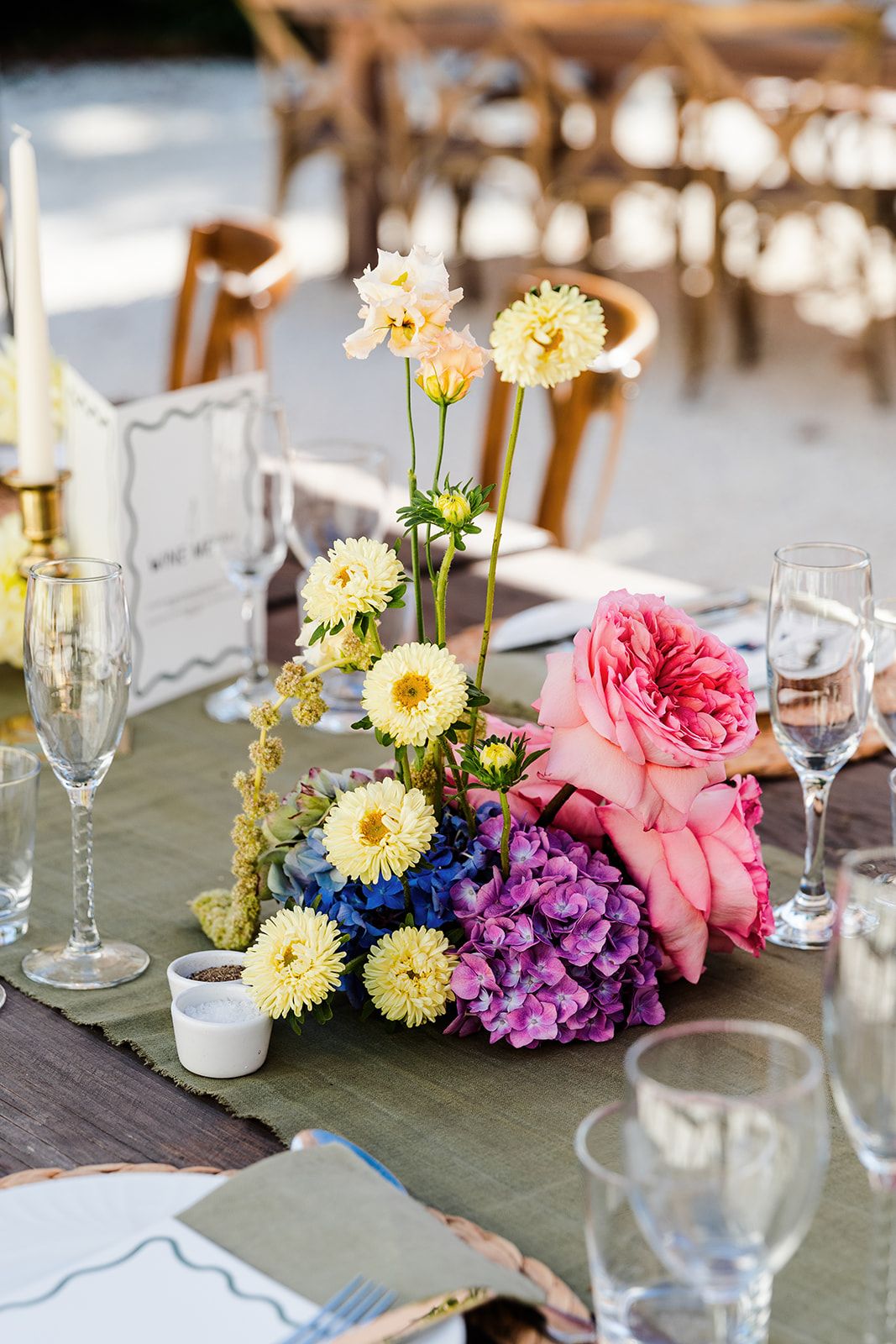 Elegant wedding table centerpiece with yellow gerbera daisies, pink garden roses and purple hydrangeas beside wine glasses.