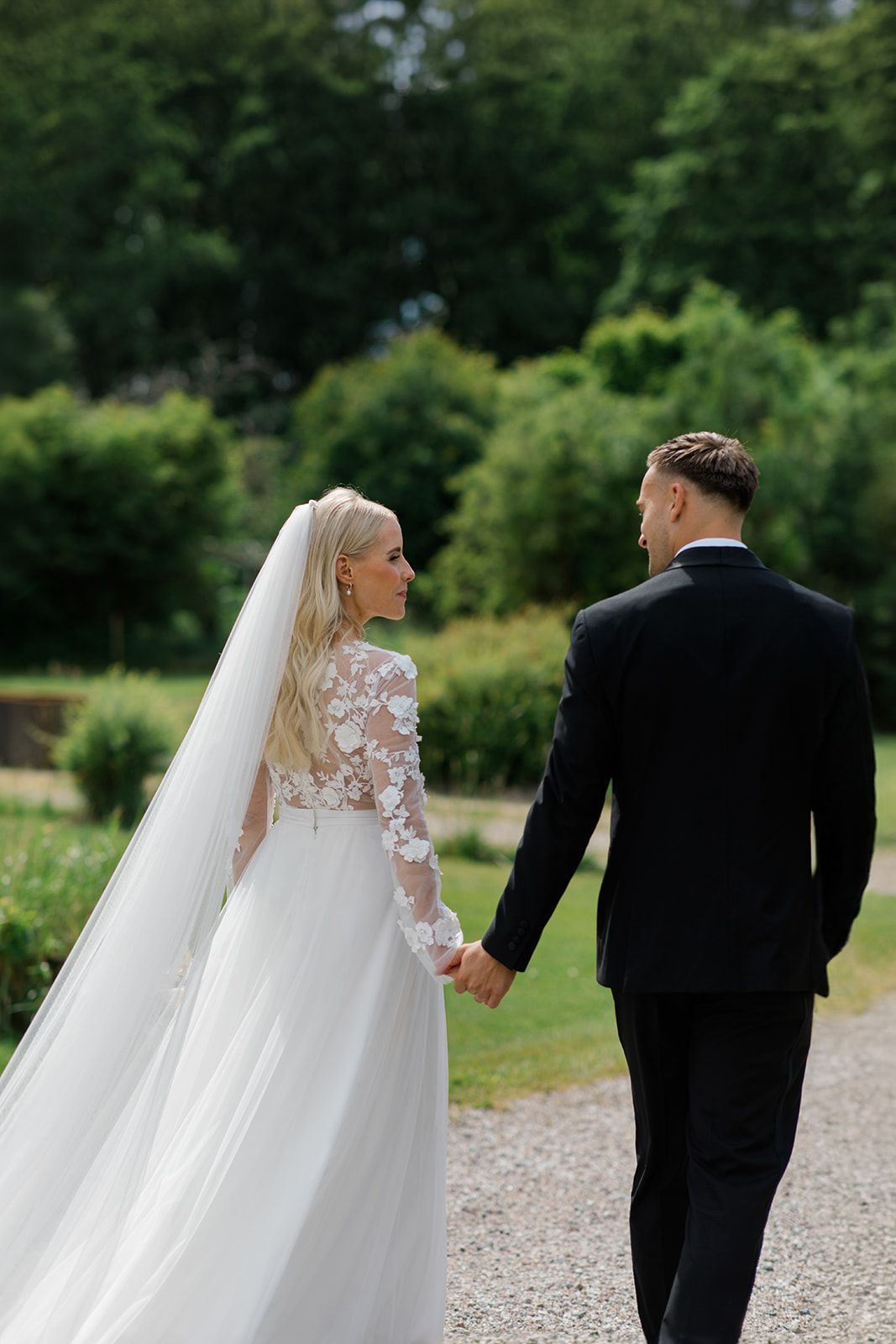 A wedding couple walks together on a gravel path surrounded by lush green trees and gardens.