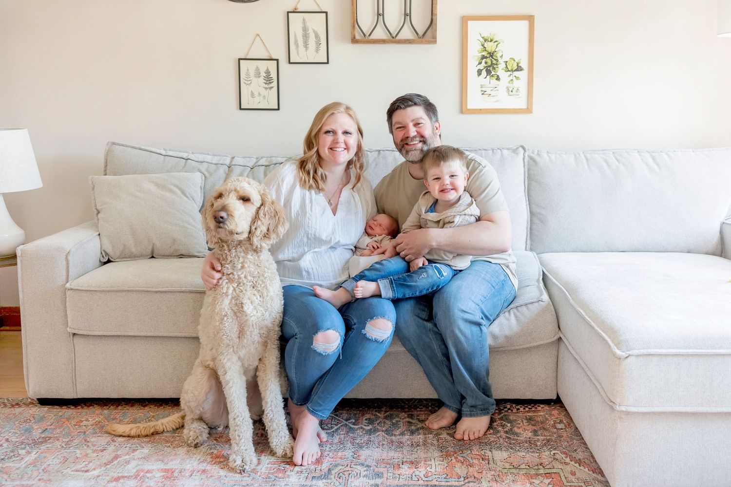In-home newborn session Clear Lake Iowa family sits together on a light gray couch in a bright living room with framed artwork on the wall.