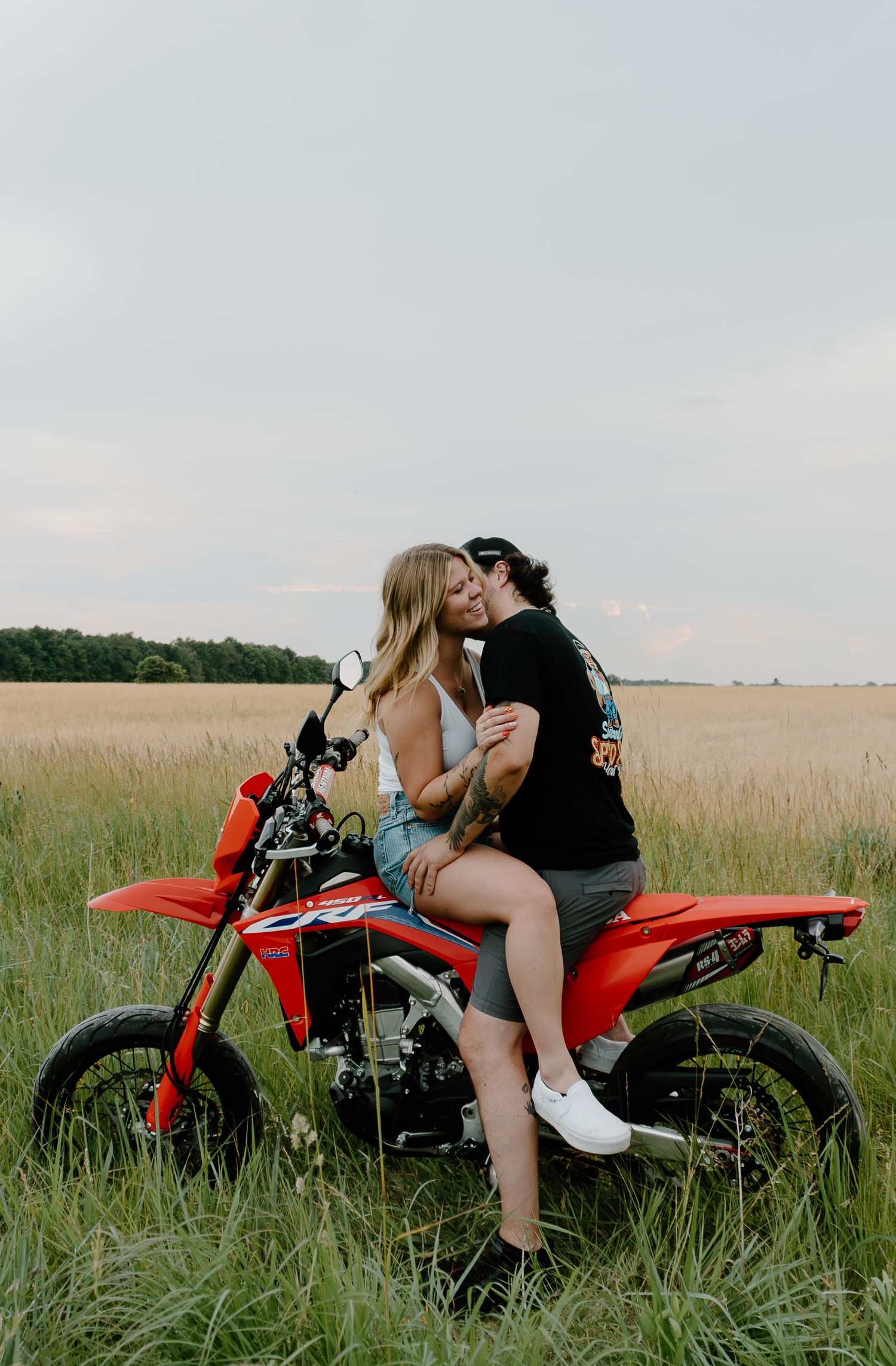 A couple shares an intimate moment on a red dirt bike in a serene grassy field during sunset.