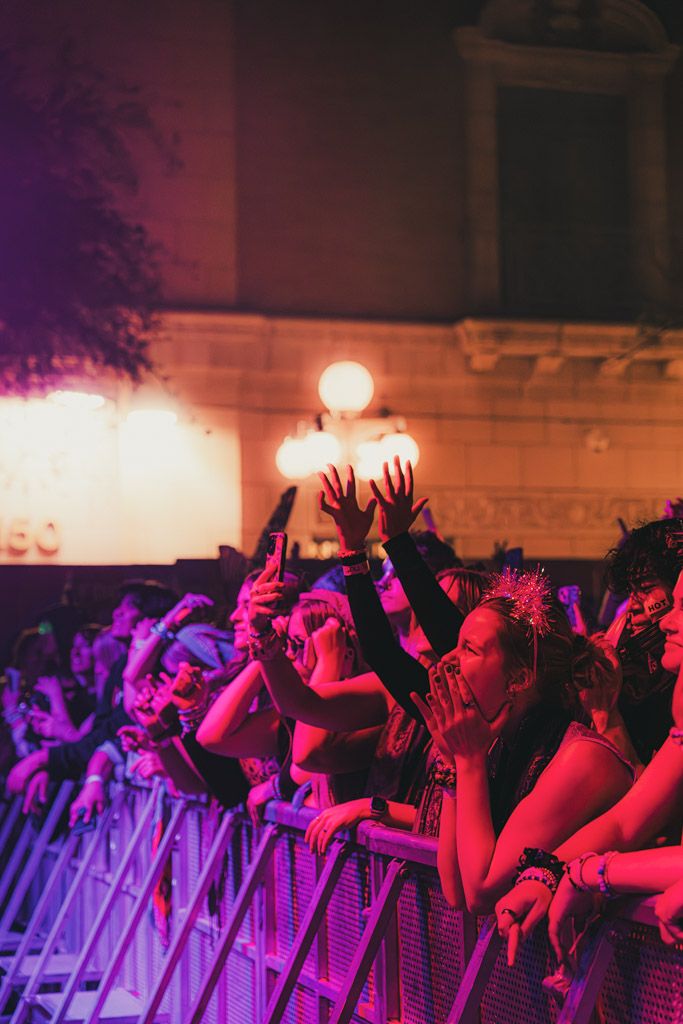 Silhouetted concert audience with raised hands against red and purple lighting at a live music show.