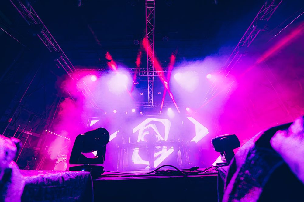 Vibrant purple and pink stage lights illuminate a concert venue with dramatic beams cutting through atmospheric haze.