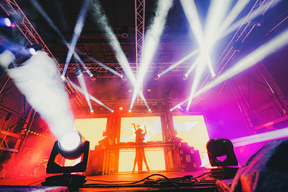 Colorful stage lighting illuminates a concert performance with dramatic beams in pink, orange and white creating an atmospheric display.