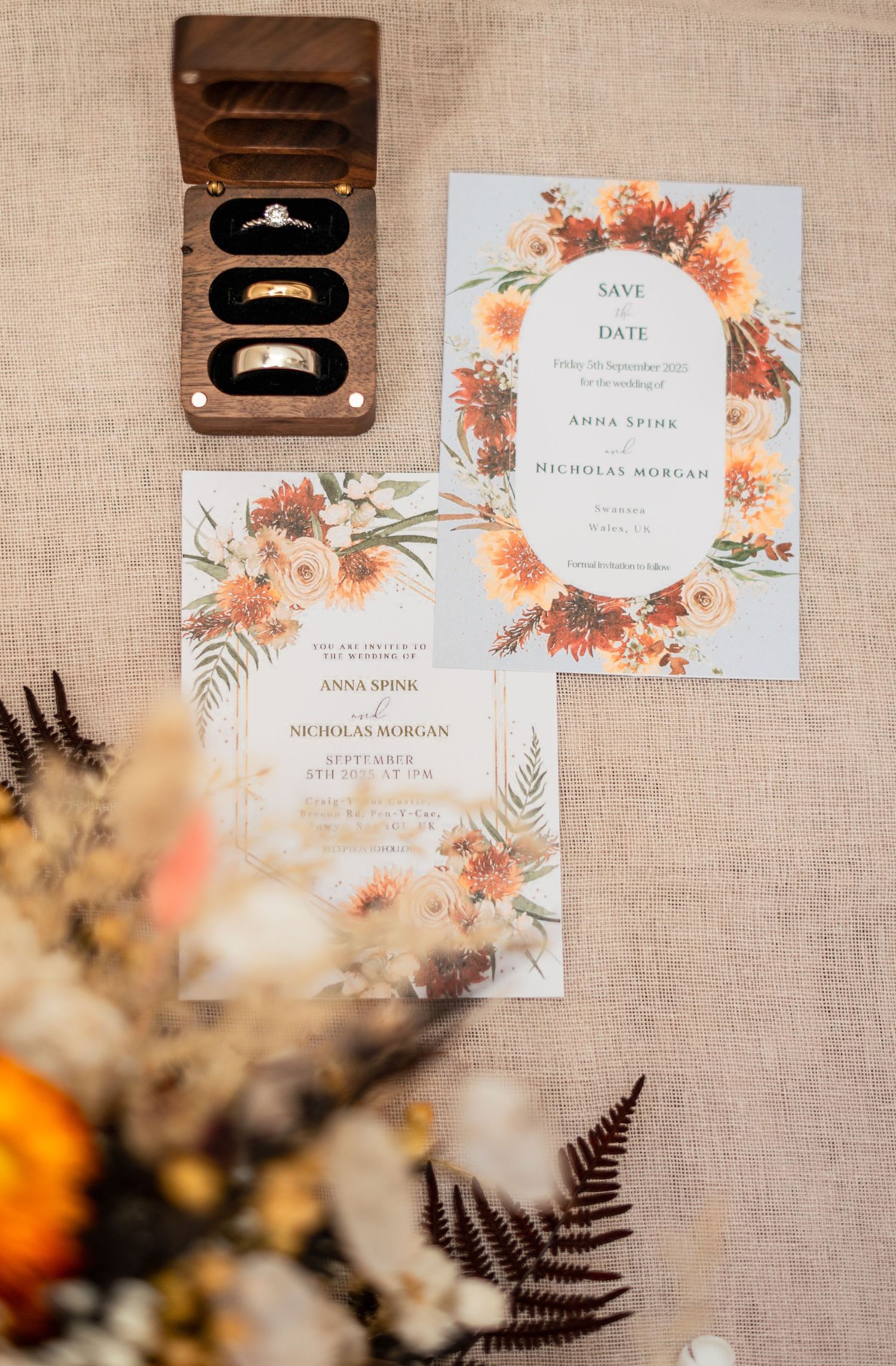 Wedding rings and floral invitation suite with orange blooms and ferns displayed on a neutral linen background.