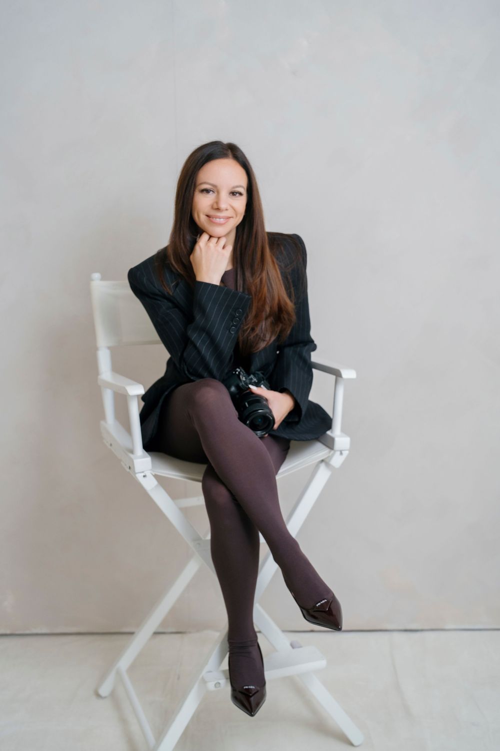 A person in a black top and brown pants poses in a white director's chair against a light background.
