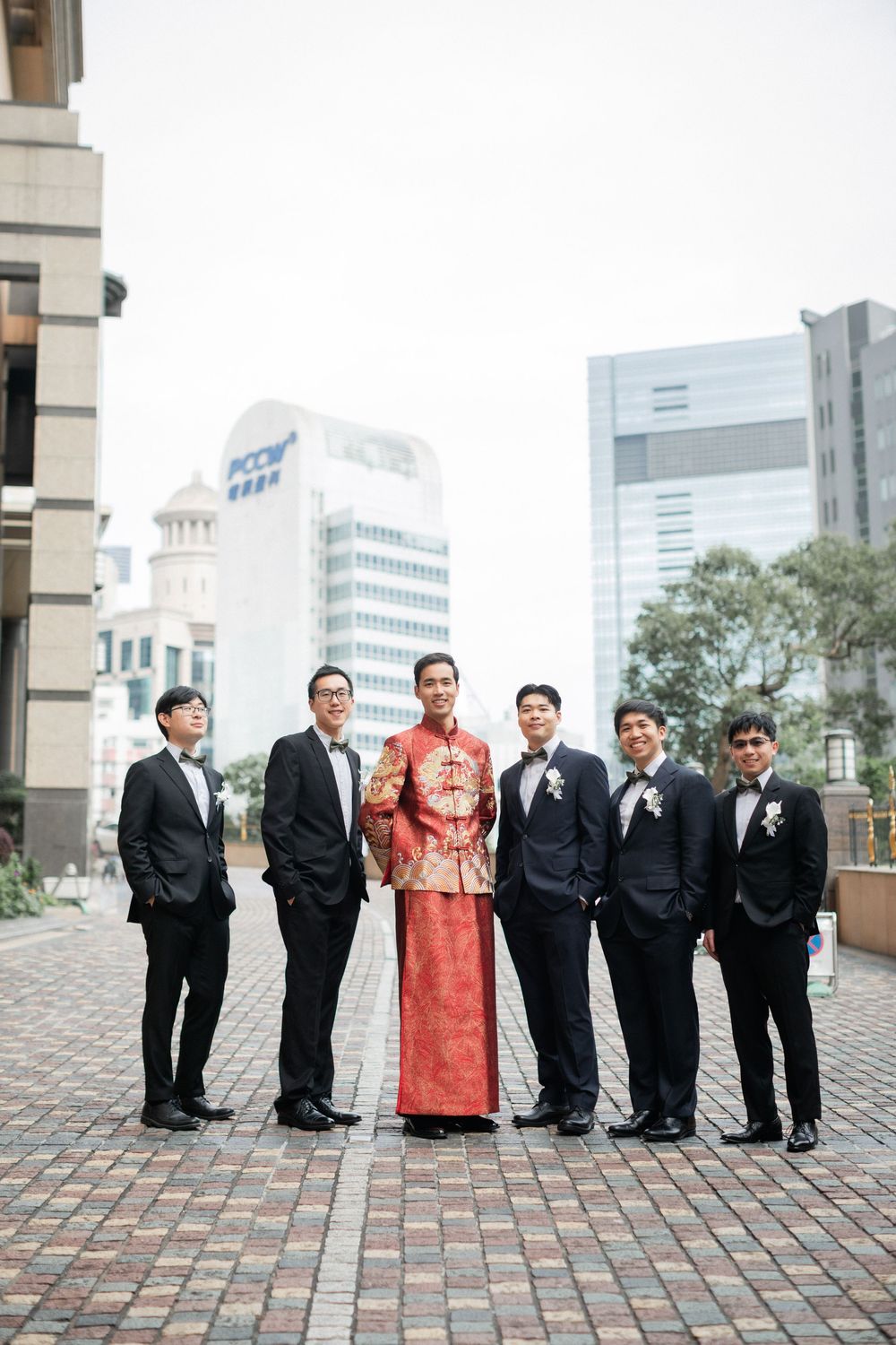 Groom in traditional red Chinese wedding attire poses with groomsmen in black suits on urban plaza.