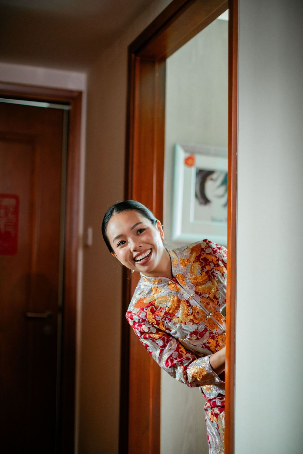 Joyful bride in traditional Chinese qipao peeking around a doorframe smiling.